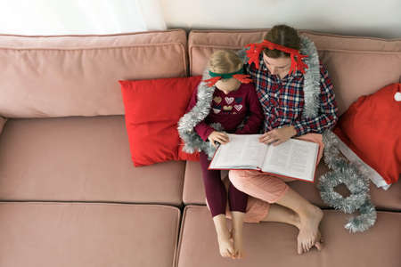 Mother with daughter wearing pajamas and antler deer horns Christmas headbands reading a book. Woman embracing her little girl while sitting on sofa on Christmasの写真素材