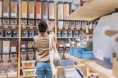 African american woman buying organic cereals and grains in sustainable zero waste grocery store. Young woman refilling reusable container in local grocery storeの写真素材