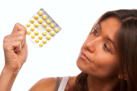 Pretty young woman looking at full medicine pack of yellow tablet pills thinking about doctor's prescription to take one or not isolated on a white backgroundの写真素材