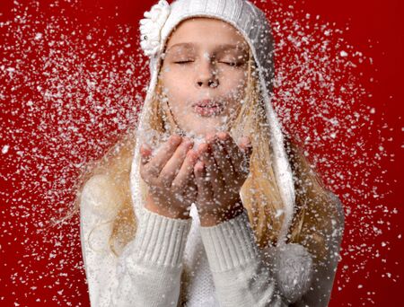 Pretty young girl in white hat blowing on the snow on her hands on red background