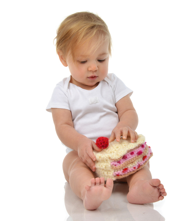 Infant child baby girl toddler sitting happy looking straight and holding piece of candy cake toy isolated on a white backgroundの写真素材
