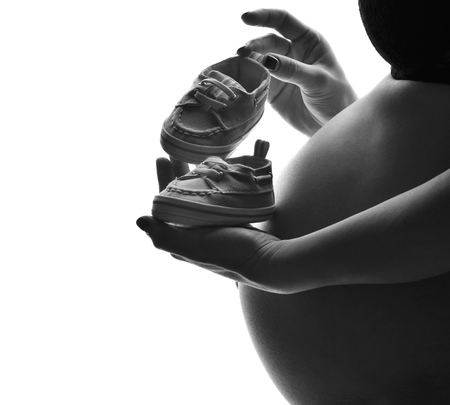 Belly of young pregnant bump woman standing with small baby boots isolated on a white backgroundの写真素材