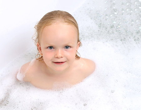 Wet baby girl toddler kid in bathing with soap suds in white bath smiling and looking at the camera