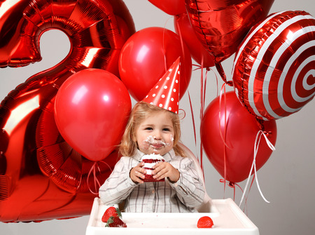 Little baby girl celebrate her second birthday with sweet cake on a light party background with red colorful balloons decorationsの写真素材