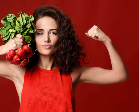 Pretty cheerful young sport woman posing with fresh radish green leaves. Dieting. Healthy eating concept on red backgroundの写真素材