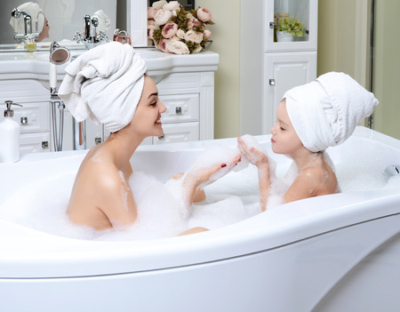 Young mother and daughter in a bathroom playing happy smiling hugging taking bath with foam