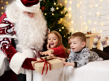 Santa Claus giving present to a happy little cute children boy and girl near Christmas tree at homeの写真素材