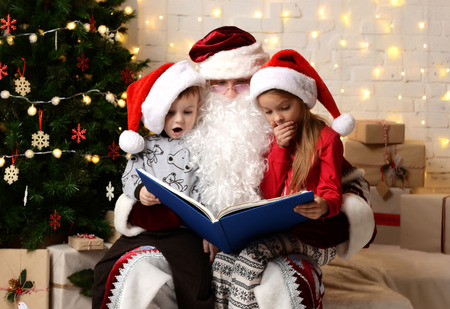 Santa Claus reading magic book to happy little cute children boy and girl kids near Christmas tree and present gift boxes at homeの写真素材