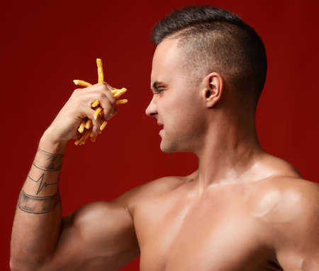 Close portrait of strong muscular athlete men hold in hand french fries unhealthy junk fast food. Unhealthy eating diet concept on dark red backgroundの写真素材