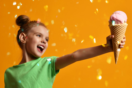 Pretty baby girl kid give away vanilla ice cream in waffles cone laughing and smiling on yellow...