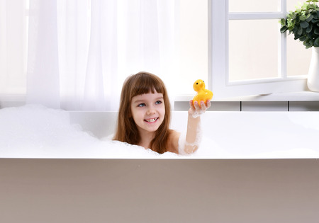 Happy little baby girl sitting in bath tub playing with yellow duck toy in the bathroom. Portrait of...