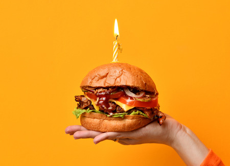 Woman hands hold big burger barbeque sandwich with beef and lit candle for birthday party on yellow backgroundの写真素材
