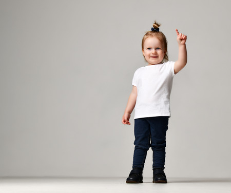 Toddler child baby girl kid standing in white free text space t-shirt pointing finger up on grey backgroundの写真素材