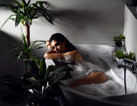 Young beautiful woman lying in bathtub and taking bath in eco bathroom with green plants