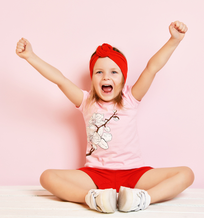 Cute baby girl kid in light pink pajamas and red headband with raised hands on pink backgroundの写真素材