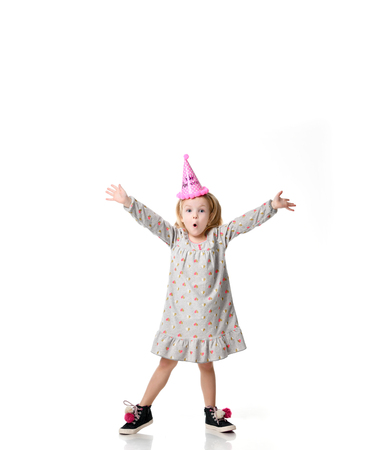 Young blond girl in birthday party princess hat surprised emotions  with hands spread up isolated on a white backgroundの写真素材