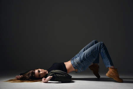 Beautiful brunette hipster woman posing in blue jeans and green jacket with windy hair posing on gray backgroundの写真素材