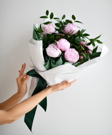 Woman hands hold bouquet of pink peony flowers with green leafs on grey backgroundの写真素材