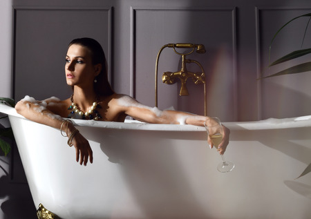 Young beautiful woman sitting in bathroom in expensive bathtub bath drinking champagne looking at the corner on dark backgroundの写真素材