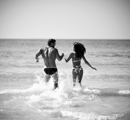 Happy cheerful couple having fun hugging running in the sea together with splashes of water on a tropical beach at sunset. Romantic vacation, honeymoon loveの写真素材