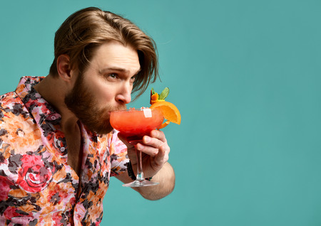 Young man in hat and sunglasses drinking red margarita cocktail drink juice with orange happy looking at the corner over blue mint backgroundの写真素材