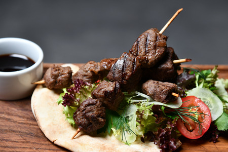 Tasty barbecue beef shashlik on wood plate with fresh vegetables and steam smoke on black backgroundの写真素材