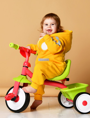 Baby girl kid riding her first bicycle tricycle in warm yellow overalls looking up on light grey backgroundの写真素材