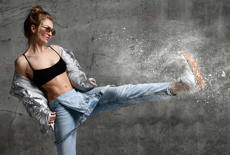 Young sport woman kick dust snow splashes with leg workout exercise stand in silver sexy thin down puffer jacket blue jeans  and sunglasses on loft wall  backgroundの写真素材
