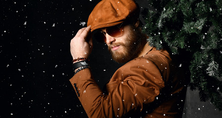 Brutal bearded man in brown leather jacket and cap hat aviator sunglasses hold Christmas tree under snowの写真素材