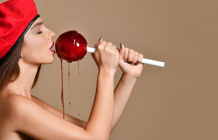 Young brunette woman in red beret hat lick huge red lollypop sweet candy with splashes on light brown backgroundの写真素材