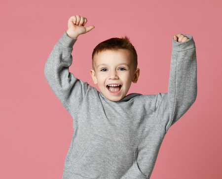 Young boy kid in grey hoodie with hands up laughing smiling isolated on pink backgroundの写真素材