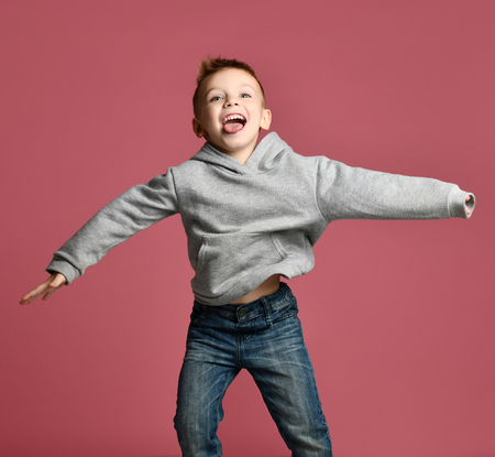 Young boy kid jumping in grey hoodie with hands spread up laughing smiling on pink backgroundの写真素材