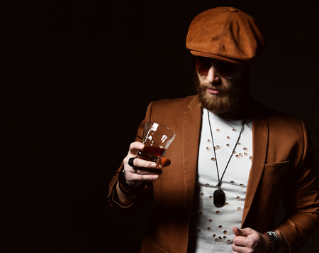 Brutal business man with beard and mustache in brown cap and jacket drink whiskey on dark backgroundの写真素材