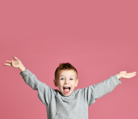 Young boy kid jumping in grey hoodie with hands spread up laughing smiling on pink backgroundの写真素材