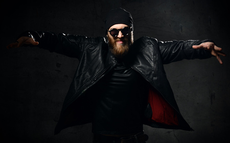 Brutal business man gangster with beard and mustache in  sunglasses  cap  and leather wear jacket with raised hands on dark backgroundの写真素材