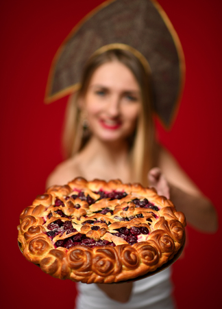 Portrait of young blonde woman in Russian traditional cap hat kokoshnik happy smiling hold sweet cherry homemade pie tasty bunの写真素材