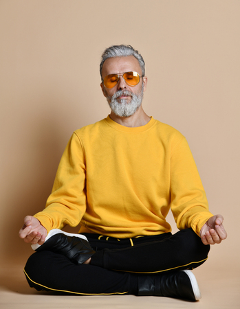 Portrait of happy senior millionaire man in yellow sunglasses stylish fashionable men practicing yoga asana stretching exercise on beige backgroundの写真素材