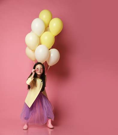 Little Korean baby girl in yellow fashion jacket and purple dress with balloons celebrate happy smiling on pink backgroundの写真素材