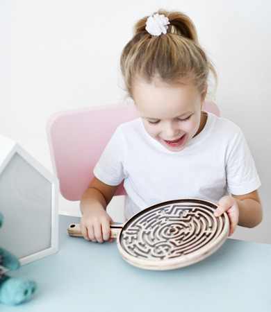 Kid girl in white t-shirt sits at the white desk and plays educational game interested with wooden labyrinth on whiteの写真素材