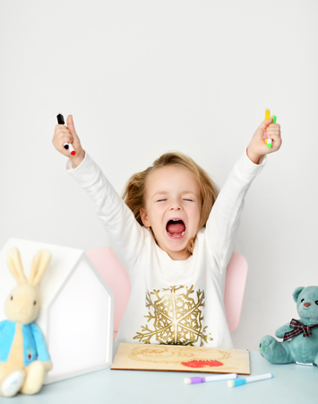 Kid girl in white shirt with thrown up hands, closed eyes and open mouth sits at the desk with ready to paint hedgehog picture grey teddy bear and rabbit on it on white backgroundの写真素材