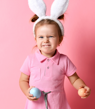 Young surprised baby girl with painted eggs wear bunny ears on Easter day. Happy Easter concept holiday celebration preparation on pink backgroundの写真素材