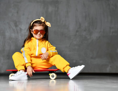 Cool kid girl in yellow hooded jumpsuit costume, sunglasses and yellow bow sits on skateboard in cool pose like just after trick on grey backgroundの写真素材
