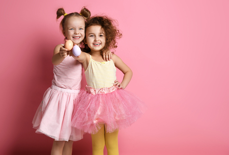 Two happy smiling kids girls demonstrate painted eggs on Easter day. Happy Easter concept holiday celebration preparation on pink backgroundの写真素材