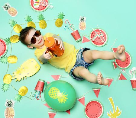 Little happy boy celebrate picnic party lying on a floor in yellow t-shirt and sunglasses happy smiling with candies and pineapple on green mint backgroundの写真素材