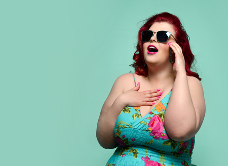 Plus-size lady overweight woman in sunglasses and colorful sundress talks on cell-phone and hear something breathtaking or incredible amazing offer isolated on mintの写真素材