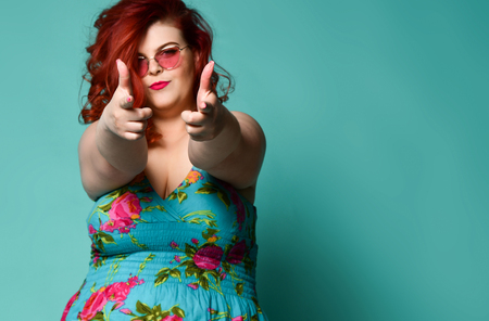 Self-confident plus-size overweight redhead lady in sunglasses and sundress shows a gesture sign finger - gun, aimed at us mint background with free text copy spaceの写真素材