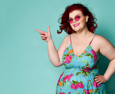 Nice smiling to us plus-size overweight lady in sunglasses and sundress point her finger at...