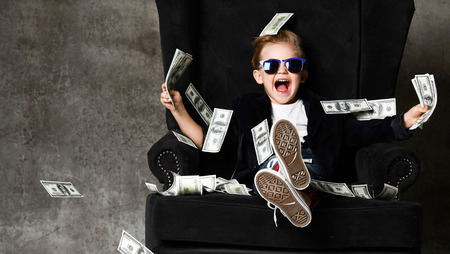 Happy young boy crazy screaming excited hold stack of dollars money. Guy celebrates success lots of money dollar bills banknotes sitting in chairの写真素材