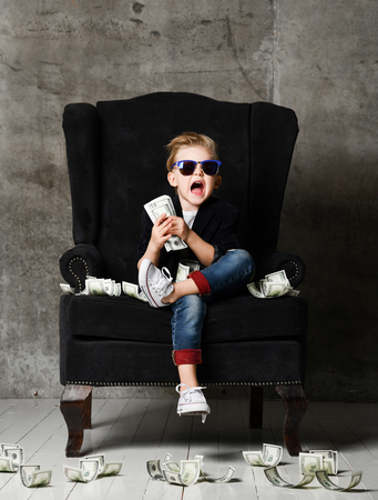 Happy young boy crazy screaming excited hold stack of dollars money. Guy celebrates success lots of money dollar bills banknotes sitting in chairの写真素材