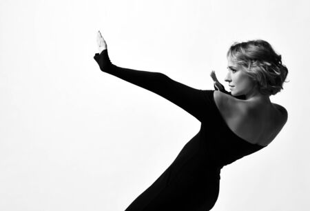 Beautiful dancer young sensual woman with short curly blonde hair in stylish black off shoulder extra long sleeve tight dress posing dancing on white backgroundの写真素材
